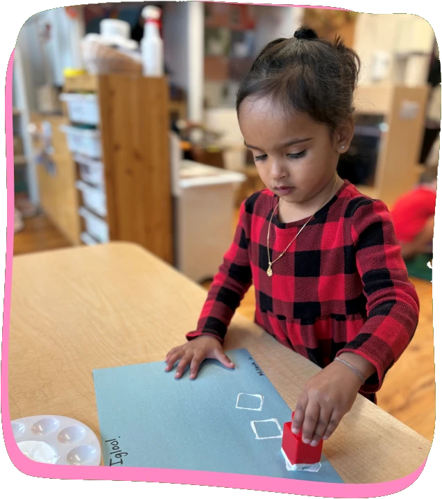 A toddler in a plaid dress stamps white shapes onto blue paper at a Songbird Academy classroom table