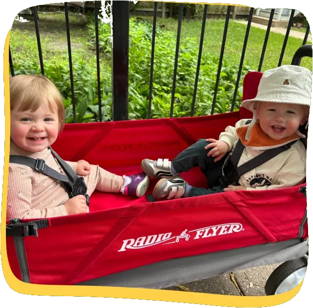 Two toddlers laughing together in a red wagon outside Songbird Academy in Chicago